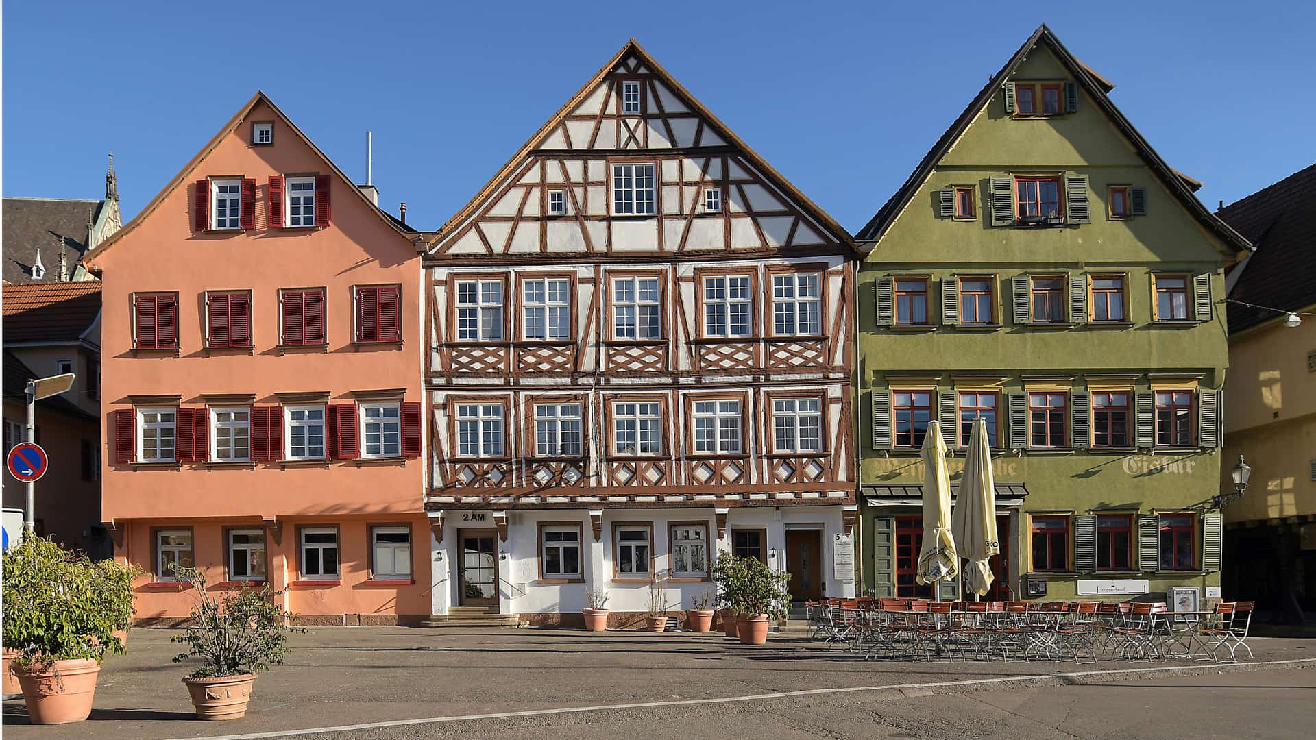 Drei Fachwerkhäuser in einem Stadtplatz, eines orange, eines mit sichtbarem Holzfachwerk und eines grün mit Fensterläden