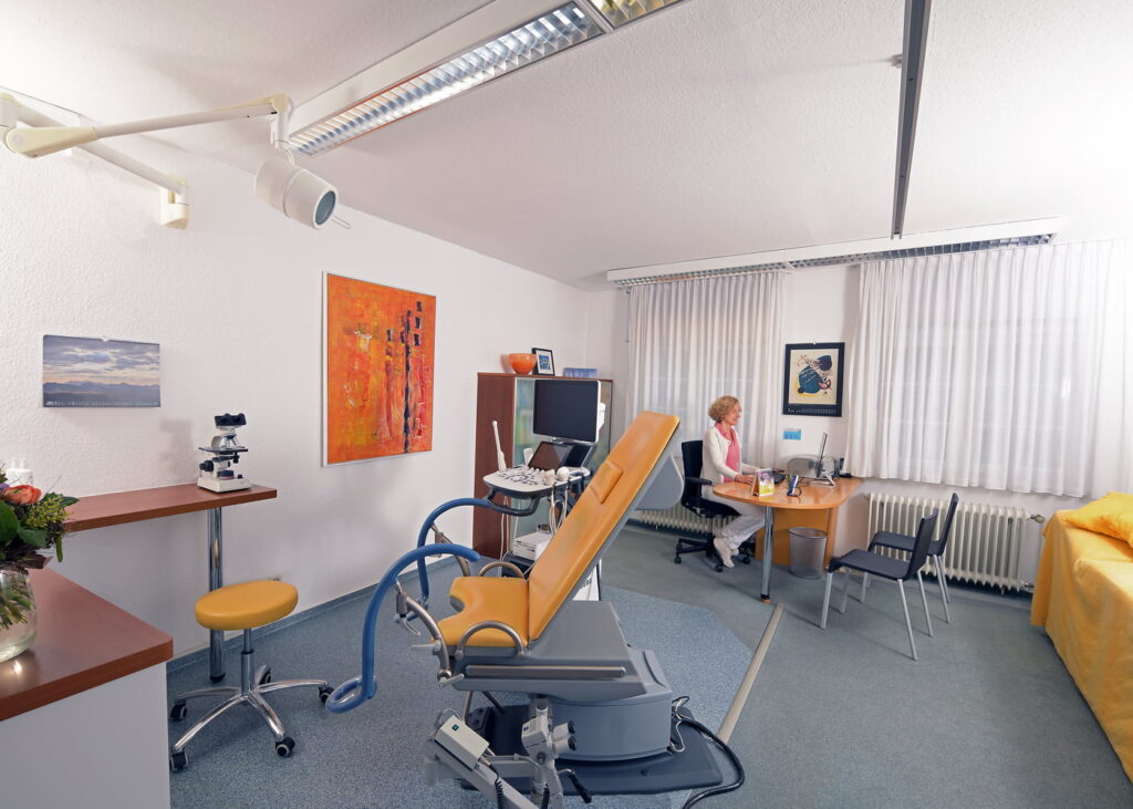 Behandlungszimmer mit gelbem Untersuchungsstuhl, Schreibtisch mit Computer, Frau sitzt am Tisch, medizinische Geräte und Kunst an den Wänden