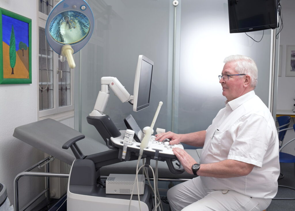 Arzt bedient Ultraschallgerät in modernem Behandlungszimmer mit Untersuchungsliege und Wandbild