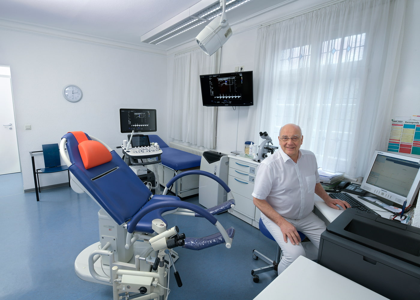 Behandlungszimmer mit gynäkologischem Untersuchungsstuhl, Monitoren und medizinischer Ausrüstung, Arzt sitzt am Schreibtisch