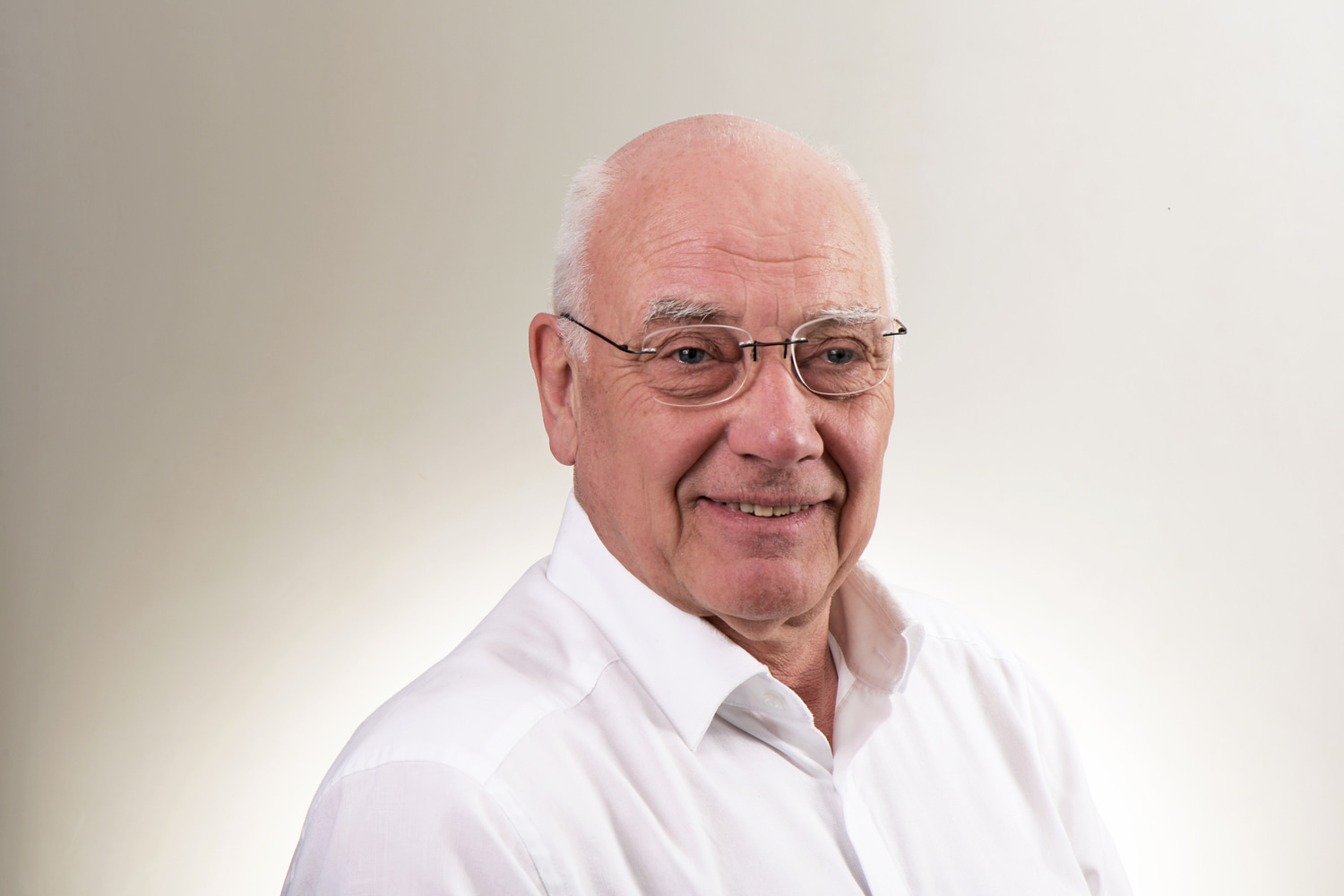 Älterer Mann mit weißem Hemd und Brille, frontal sitzend vor hellem Hintergrund