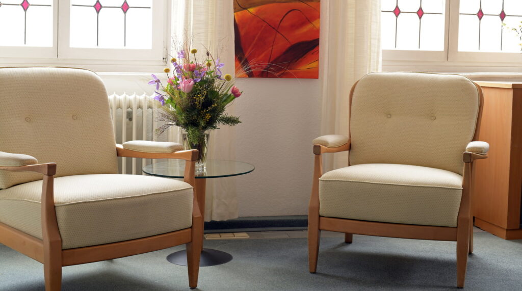 Zwei beige Sessel mit Holzrahmen stehen in einem hellen Raum neben einem Glas-Beistelltisch mit Blumenstrauß