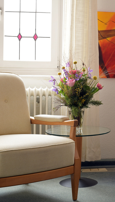 Beiger Sessel mit Holzrahmen neben rundem Glastisch mit Vase voller bunter Blumen vor Fenster mit weißem Vorhang