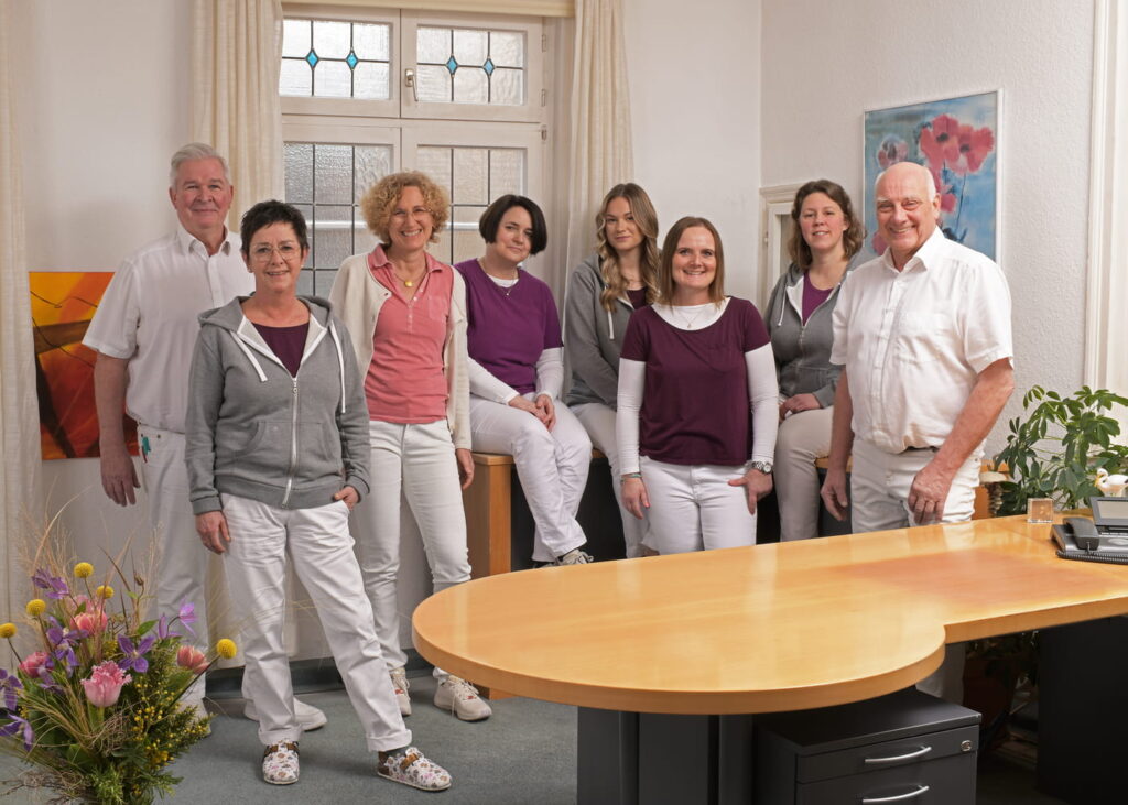 Acht Personen in weißer und bunter Arbeitskleidung stehen und sitzen in einem hellen Büro mit großem Holztisch und Blumenarrangement